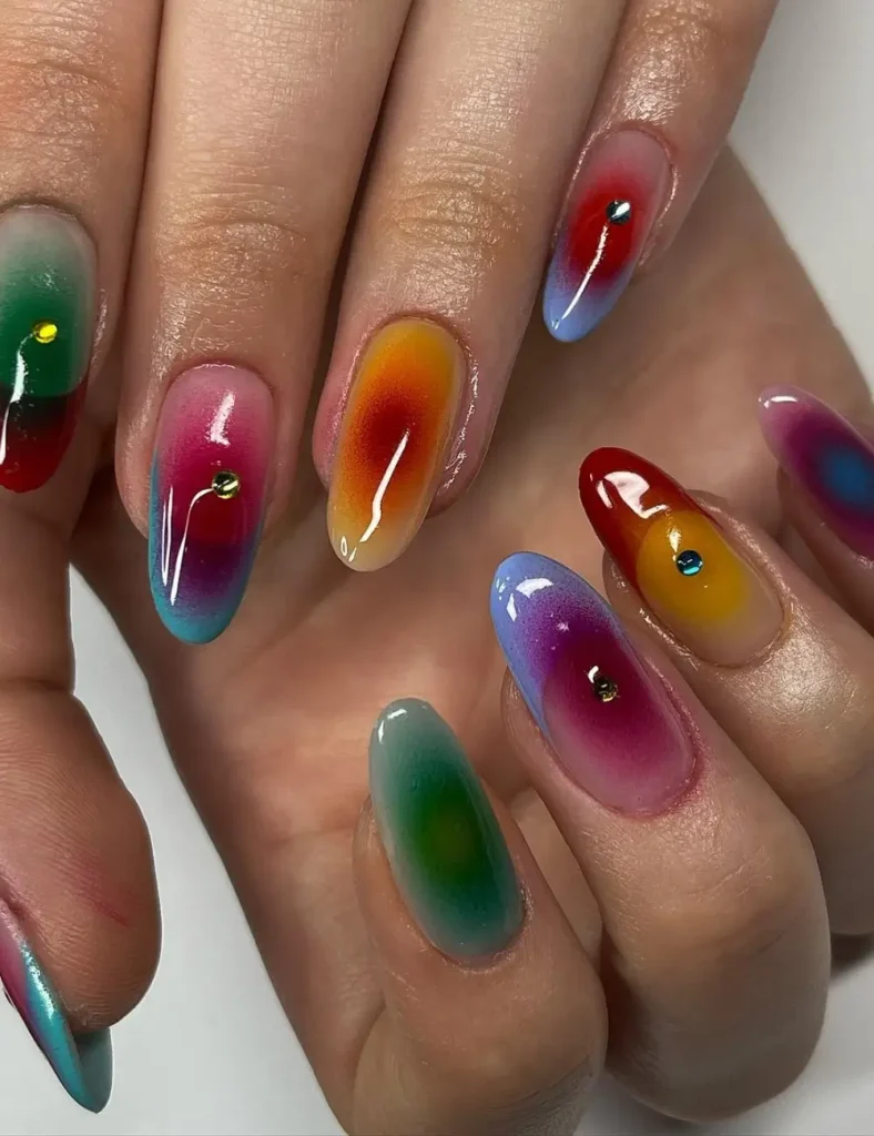 Almond nails on a sheer base with mismatched jewel-tone aura glows in green, red, amber, magenta, and blue, each with a small gem accent