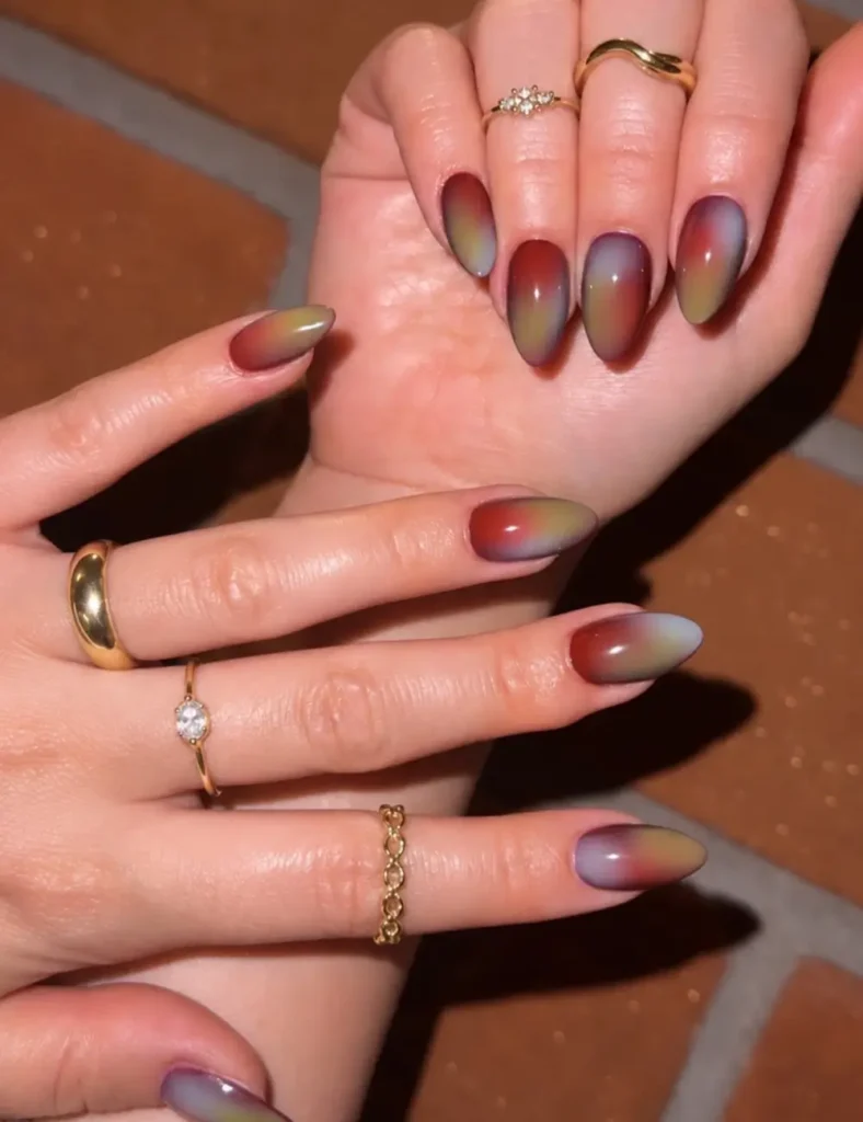 Almond nails with a blended aura of rust red, mustard yellow, and soft lilac gray, worn with stacked gold rings
