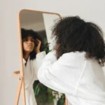 Woman with natural curly hair in a white robe checking her curls in a wooden floor mirror, showing how to moisturize low porosity hair
