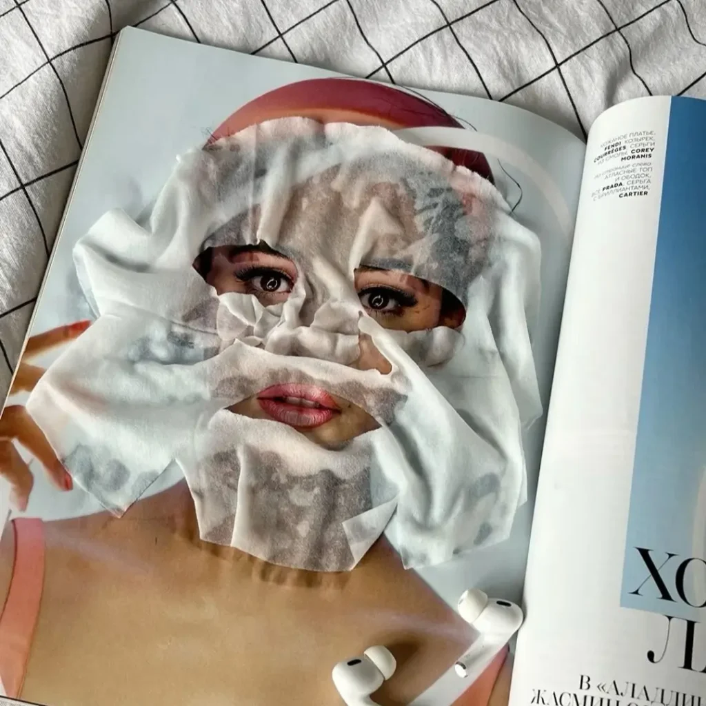 White sheet mask laid over a model’s face on an open magazine page, resting on a gray checked bedspread beside a pair of white wireless earbuds.
