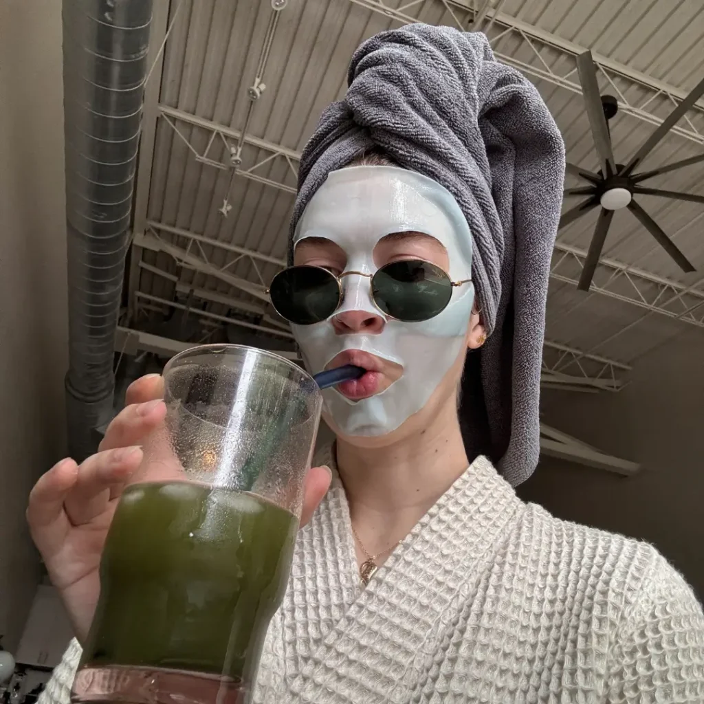 Woman in a white sheet mask, round sunglasses, gray towel hair wrap, and waffle-knit robe sipping a green drink through a straw in a selfie.