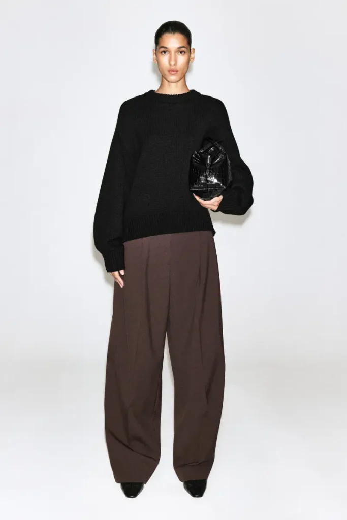 Woman wearing COS dark brown relaxed wide-leg pants with front pleats, styled with a black ribbed crewneck sweater and a black croc-effect handbag