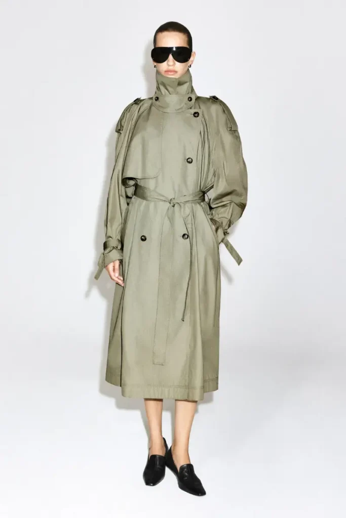 Woman wearing a COS olive green cotton trench coat with a mock-neck collar, double-breasted front, and tie belt, styled with black loafers and sunglasses