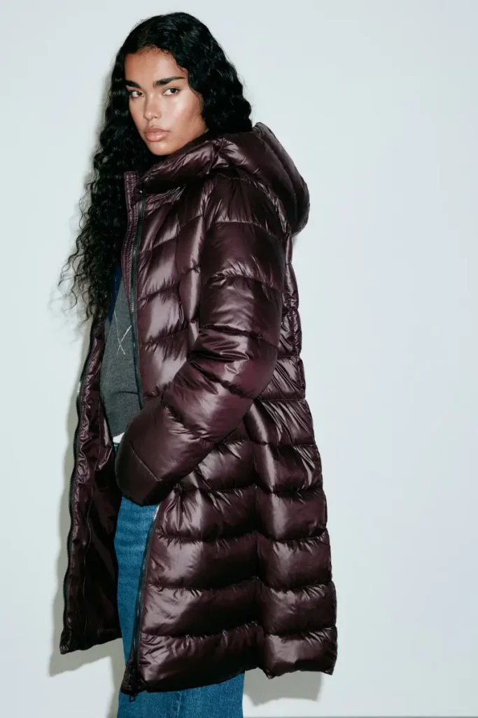 Woman wearing a Mango glossy burgundy midi quilted anorak with a hood and high padded collar, styled with blue pants