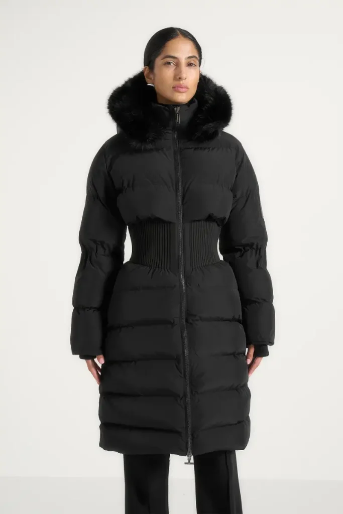 Woman wearing a Maniรจre De Voir black longline puffer coat with a faux fur trimmed hood and ribbed corset waist panel