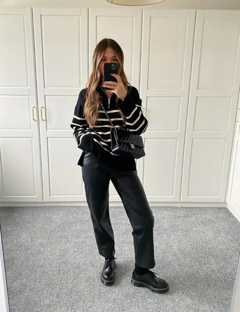 Woman wearing a black-and-cream striped sweater, black leather-look trousers, chunky black lace-up boots, and a black shoulder bag in front of white paneled doors
