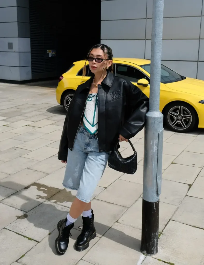Woman wearing a black leather jacket over a light top, a light blue denim midi skirt, black socks, chunky black lace-up boots, and sunglasses on a city sidewalk