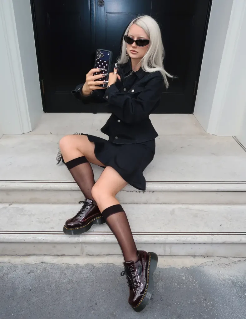 Woman wearing a black mini dress, sheer black tights, black lace-up Dr. Martens boots, and sunglasses while sitting on outdoor stone steps