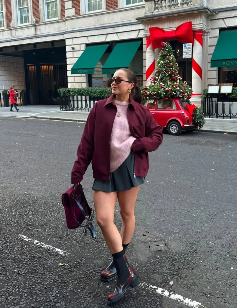 Woman wearing a burgundy blazer over a pink shirt, a dark pleated mini skirt, sheer tights, black lace-up boots, and carrying a burgundy handbag on a city street
