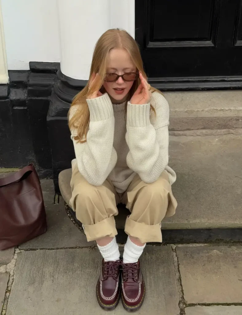 Woman wearing a chunky cream sweater, beige pants, white socks, burgundy lace-up Dr. Martens boots, and sunglasses while sitting on stone steps