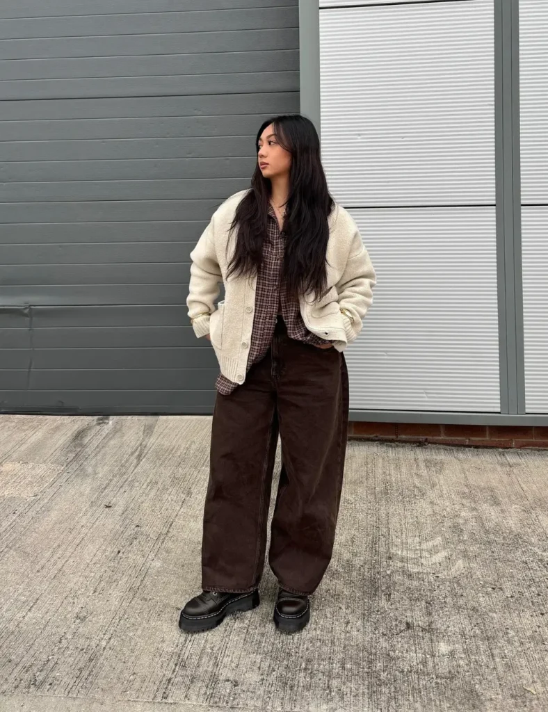 Woman wearing a cream jacket, relaxed dark brown pants, and chunky black lace-up boots while standing in front of a gray metal garage door