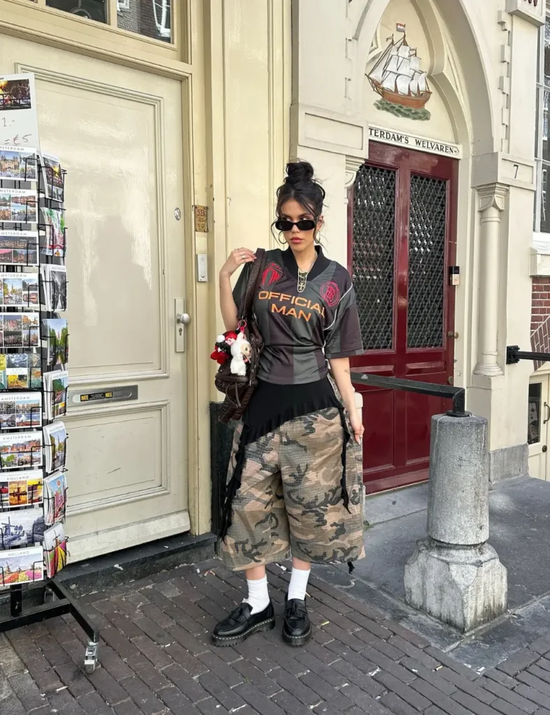 Woman wearing a dark graphic T-shirt, a long camouflage skirt, black lace-up boots, sunglasses, and a small shoulder bag while standing on a city sidewalk