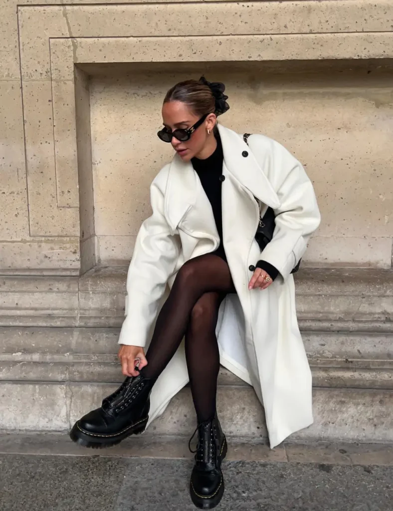 Woman wearing a long cream trench coat over a black outfit, sheer black tights, chunky black lace-up boots, and sunglasses while sitting on stone steps