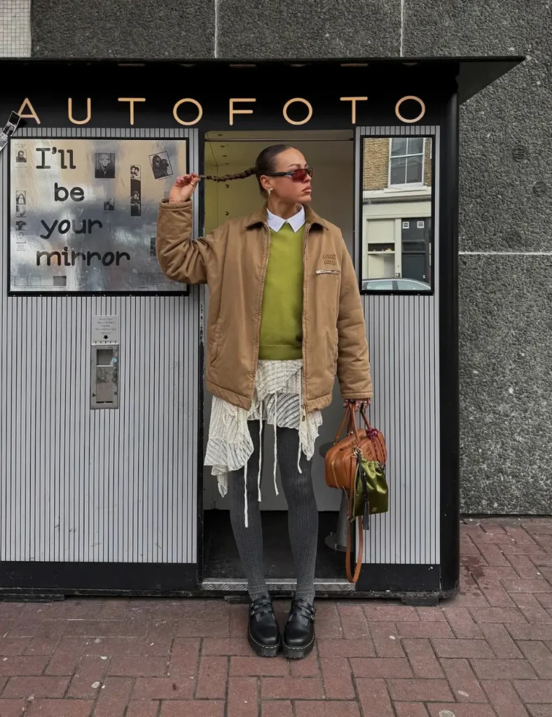 Woman wearing a tan jacket over an olive-green sweater, a gray scarf, black pants, chunky black lace-up boots, and sunglasses while standing outside a storefront