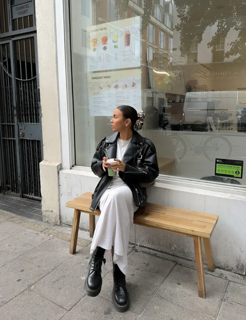 Woman wearing a white midi dress, a black leather jacket, and chunky black lace-up boots while standing near a wooden bench outside a storefront