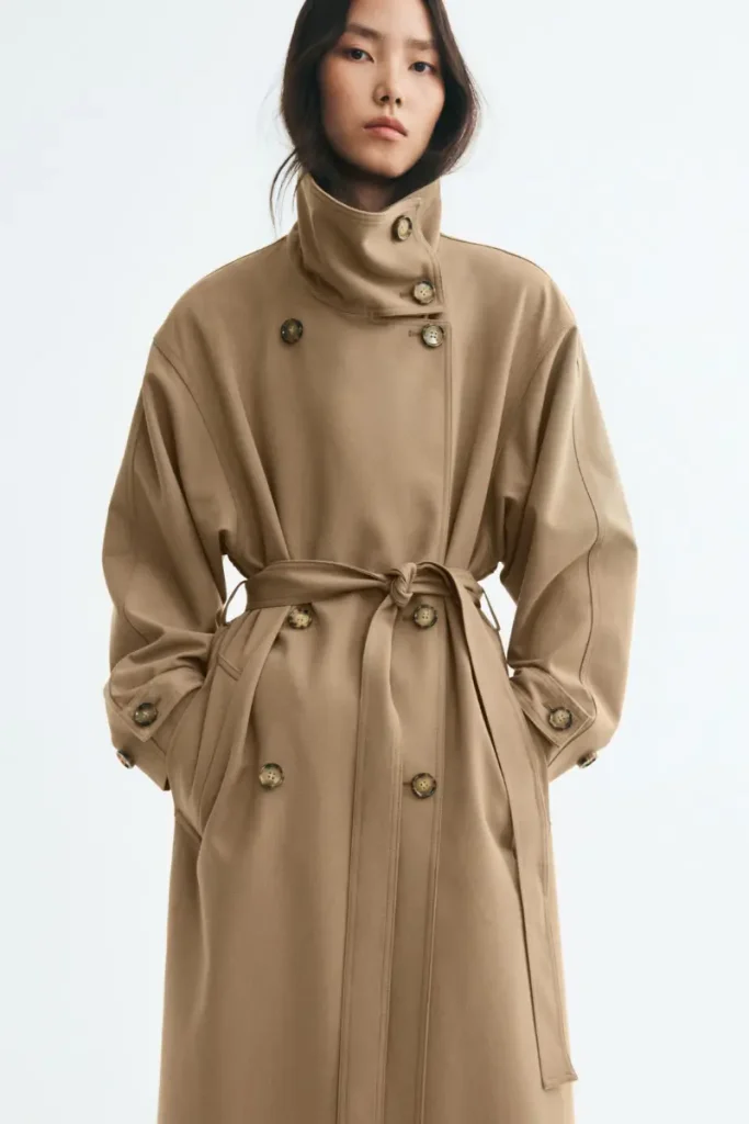 Woman wearing an H&M oversized beige twill trench coat with a high funnel collar, double-breasted tortoiseshell buttons, and dropped shoulders, styled with pinstriped pants