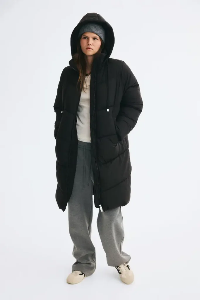 Woman wearing an H&M oversized black knee-length puffer coat in quilted nylon with a drawstring hood pulled up over a gray beanie, styled with gray sweatpants