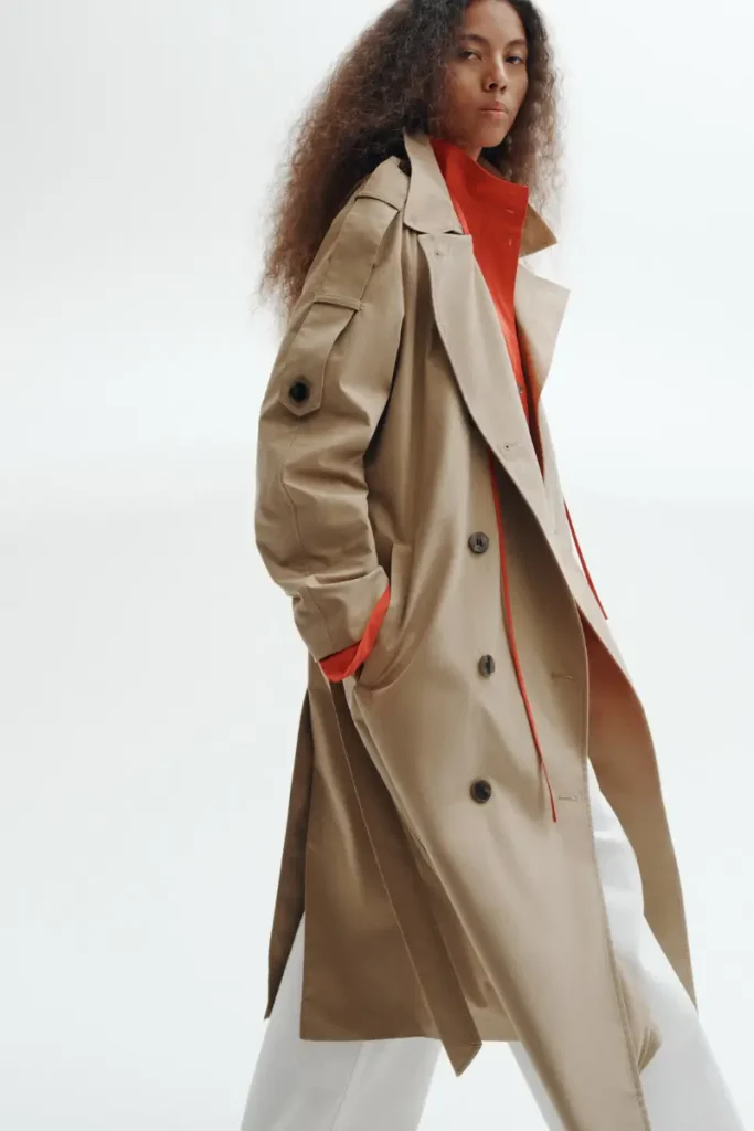 Woman wearing an H&M oversized dark beige maxi trench coat with wide notched lapels and double-breasted buttons, styled over a red shirt and white pants