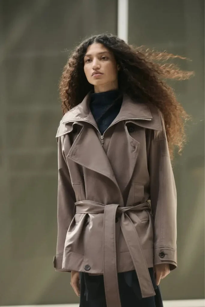 Woman wearing an H&M short oversized dark taupe trench coat with a zip-up funnel collar, notched lapels, and a tie belt, styled with a dark midi skirt and loafers