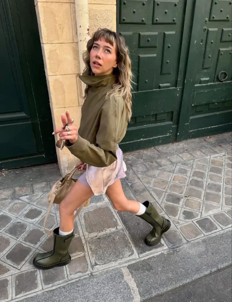 Woman wearing an oversized olive-green jacket, pale pink shorts, white socks, and chunky olive-green lace-up boots while walking along a cobblestone street